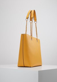 Żółta skórzana torba typu tote o eleganckim wzornictwie, z uchwytami ozdobionymi łańcuszkami i kontrastowym przeszyciem wzdłuż krawędzi.