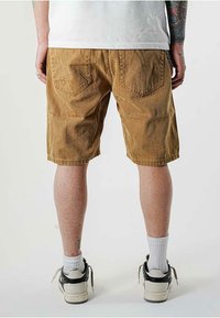 Pantaloni corti di velluto a coste marroni con una superficie testurizzata, tasche posteriori e orlo al ginocchio; indossati con calze bianche e sneakers in bianco e nero.