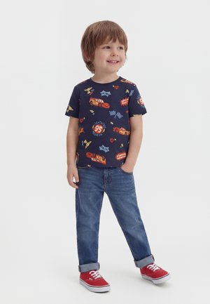 Νεαρός αγόρι που χαμογελά, φορώντας μπλε σκούρο μπλουζάκι με στάμπα Lightning McQueen, μπλε τζιν με γυρισμένα μανίκια και κόκκινα αθλητικά παπούτσια.