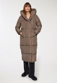 Lunga giacca trapuntata in taupe con un ampio cappuccio, tasche laterali e taglio dritto. Indossata con jeans scuri e stivali neri robusti.
