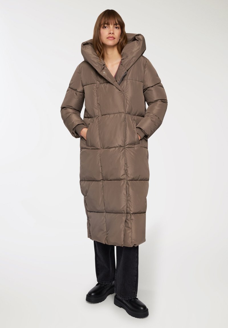 Lunga giacca trapuntata in taupe con un ampio cappuccio, tasche laterali e taglio dritto. Indossata con jeans scuri e stivali neri robusti.
