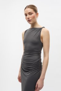 Femeie cu părul tras pe spate poartă o rochie gri închis fără mâneci, cu pliuri, stând într-un fundal alb simplu, cu o expresie neutră.