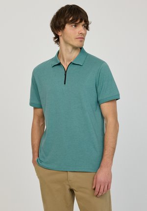 Homme portant un polo à manches courtes bleu sarcelle avec une fermeture éclair noire, pantalon beige, regardant vers la droite, debout devant un fond clair uni.