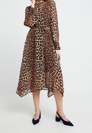 Kobieta w długim rękawie, z paskiem, w midi sukience w zwierzęcy print, połączonej z granatowymi sandałami na obcasie, stojąca na tle białym.