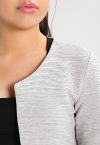 Šedý svetr s texturou a vroubkovaným materiálem, s kulatým výstřihem, přes černý top. Detailní záběr na oblast límce a horní část hrudníku.