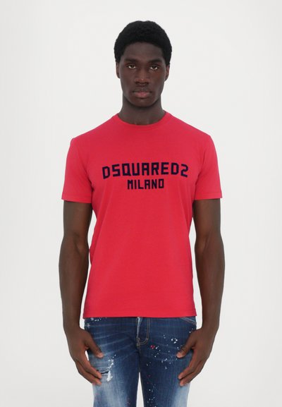 T-shirt rossa con testo "DSQUARED2 MILANO" in blu navy, maniche corte, colletto tondo. Indossata con jeans in denim blu strappati con macchie di vernice.