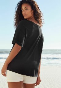 Zwarte oversized shirt met opgerolde mouwen, met een losse pasvorm en een versleten zoom, gedragen met witte shorts tegen een strandachtergrond.