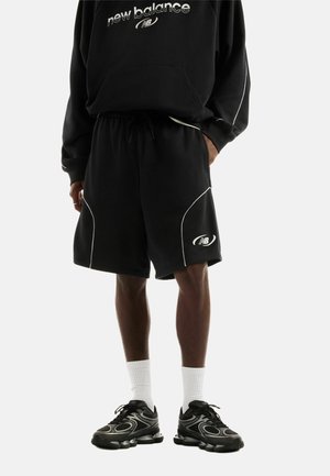 Person trägt schwarzen New Balance Sweatshirt und passende schwarze Shorts mit weißen Paspeln, schwarze Sneakers und weiße Sportsocken, stehend.