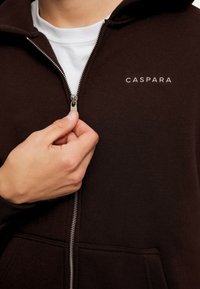 Brun zip-up hettegenser med hvit logo "CASPARA" på brystet. Har metallglidelås og frontlommer. Myk, teksturert stoff.