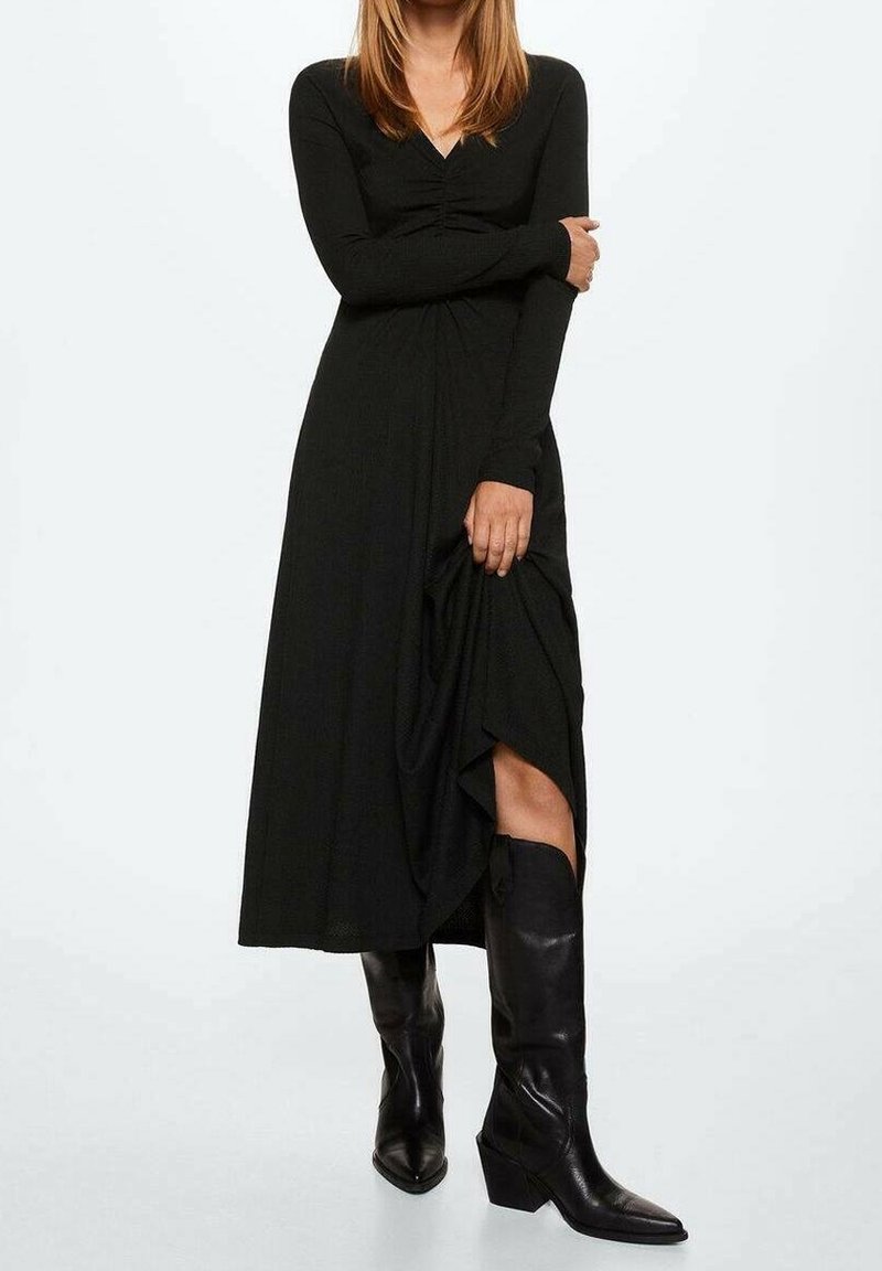 Černé dlouhé šaty s výstřihem do V, úzkou korzetovou částí a splývavou sukní, která má boční rozparek. Vpředu kombinováno s černými koženými botami do kolen.