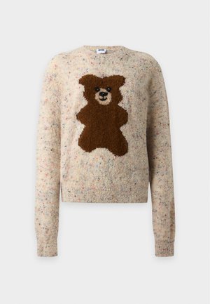 Femme portant un pull beige oversize avec un motif ours en peluche marron et un jean large bleu clair, debout sur un fond uni.