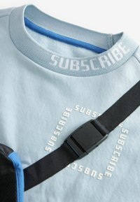 Gaiši zils kokvilnas sporta krekls ar ribotu apkakli, baltiem vārdiem "SUBSCRIBE" un melnu regulējamu siksnu ar plastmasas sprādzi.