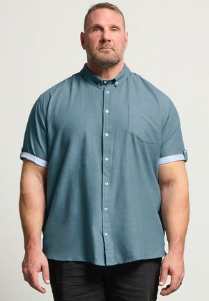 Mann mit Bart, der ein türkisfarbenes kurzärmeliges Hemd mit Knöpfen und schwarze Hose trägt, steht vor einem einfarbigen hellen Hintergrund und schaut nach vorne.