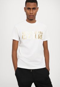 Balta medvilnė T-marška su auksiniu metaliniu užrašu "VERSACE JEANS COUTURE". Klasikinis apvalus kaklas ir trumpomis rankovėmis, su laisvu siluetu.