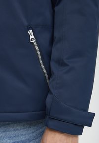 Navy jacket featuring a zippered pocket and cuff detail, made from a smooth, waterproof material with a sleek, structured design.