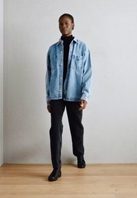 Chemise en denim bleu clair avec des boutons, portée sur un col roulé noir, associée à un pantalon sombre et des bottes noires à lacets. Texture lisse, coupe décontractée.