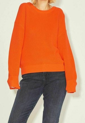 Person iført en lys orange, langærmet strikket trøje kombineret med mørkeblå jeans mod en neutral baggrund.