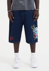 Tummansiniset farkkushortsit, joissa on sinisen, punaisen ja vihreän värinen brodeerattu lohikäärme. Vasemmassa jalassa on punainen logoyksityiskohta.