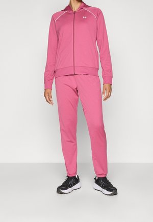 Persoon draagt een roze hoodie met rits en bijpassende joggingbroek met zwart-witte sportschoenen, staand tegen een effen achtergrond.