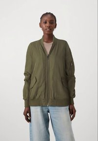 Mulher em pé a usar uma jaqueta bomber de fecho em verde oliva, uma blusa bege, calças largas de ganga azul claro e brincos de argola, contra um fundo simples.