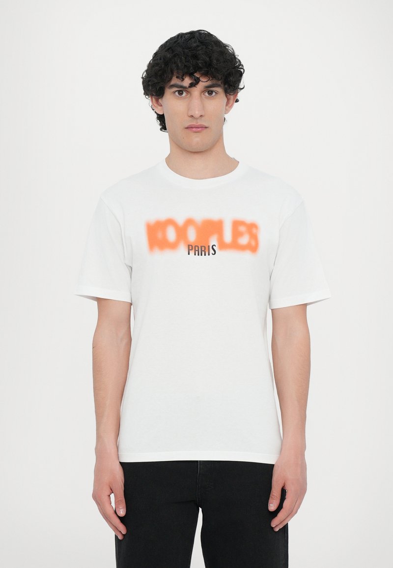 T-shirt en coton blanc à manches courtes avec le texte noir "100 PLES" et un effet de spray orange, stylisé avec un col rond.