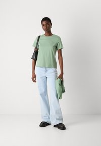 T-shirt di cotone verde chiaro con maniche corte e un piccolo design ricamato, abbinato a jeans svasati blu chiaro e scarpe nere.