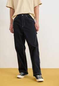 Pantalons noirs amples en tissu durable avec des coutures blanches contrastantes. Assortis à des baskets en toile noires et une chemise beige.