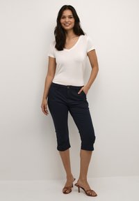 Balta V formos iškirptė marškinėliai derinami su tamsiai mėlynomis iki kulkšnių kelnių, turinčiomis priekinę kišenę ir sagos detalę. Rudos, juostelės aukštakulniai sandalai užbaigia aprangą.