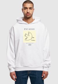 Hvid hættetrøje med en frontgrafik af en due, tekst som beskriver "Picasso" og "Fredens Due," med en lomme. Almindelig pasform.