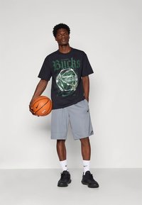 Camiseta negra con gráfico y logo verde de "Bucks", pantalones cortos deportivos grises, zapatillas negras, sosteniendo un baloncesto. Fondo sencillo.