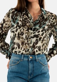 Transparante button-up blouse met een luipaardprintpatroon in zwart, bruin en teal tinten, gedragen met high-waisted blauwe denim jeans.
