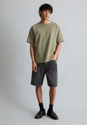 Homme debout devant un fond gris, portant un haut à manches courtes texturé olive, un short en jean noir, des chaussettes noires et des mocassins noirs.