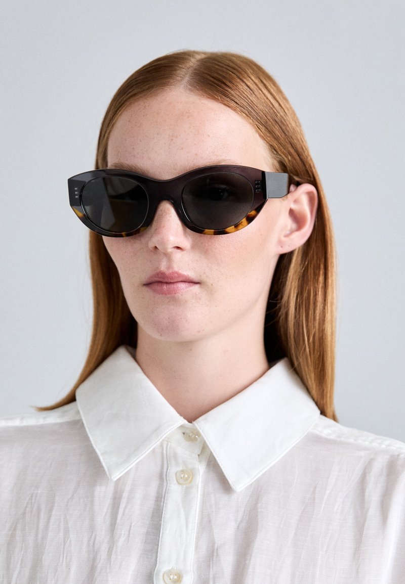 Black and tortoiseshell sunglasses with a sharp cat-eye shape, dark lenses, and geometric accents on the temples; lightweight material.