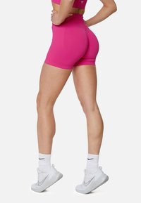 Táhnuté ružové športové šortky s vysokým pásom, vyrobené z hladkej a pružnej látky. Nosené s bielymi teniskami a členkovými ponožkami.