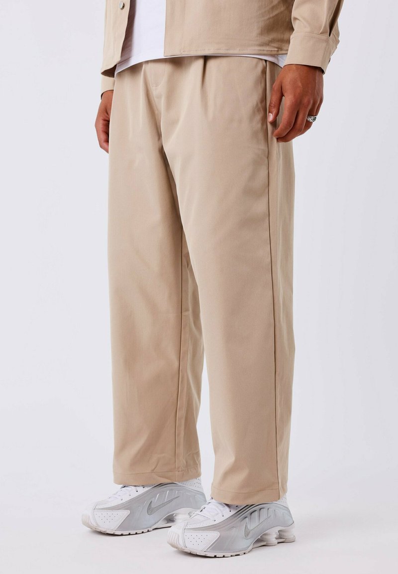 Beige broek met een relaxed fit, met een gladde textuur en een geplooide voorkant, gecombineerd met witte en zilveren sportieve schoenen.