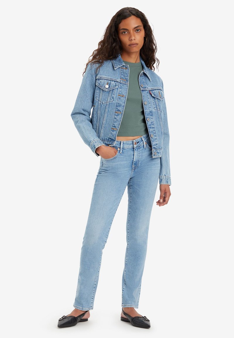 Chaqueta de denim azul claro con dos bolsillos frontales, combinada con una blusa verde y jeans azules. Unos zapatos planos negros completan el conjunto.