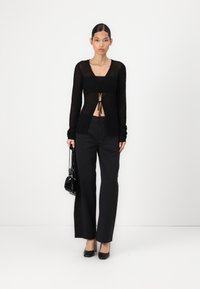 Cardigan nero a maglia con una cintura davanti, abbinato a pantaloni neri a vita alta. Il modello tiene una piccola borsa nera, indossando tacchi neri.