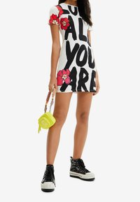 Krátké bílé šaty s výrazným černým textem a červenými květinovými vzory; doplněné žlutou taškou a vzorovanými černými vysokými teniskami.