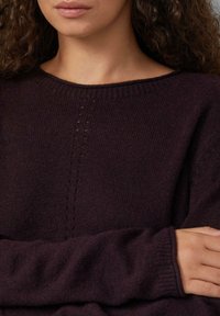 Vrouw met krullend haar in een donkerpaarse gebreide trui met een subtiel verticaal patroon, met gekruiste armen, afgeknipt onder de ogen.