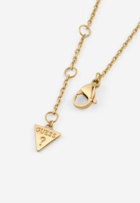 Goudkleurige ketting met een driehoekig hangertje gegraveerd met "GUESS?" en een karabijnsluiting. Eenvoudig ontwerp en gepolijste afwerking.