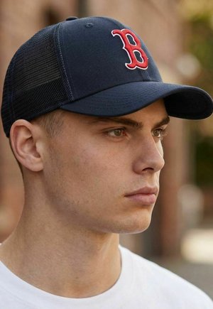 Junger Mann, der eine schwarze Baseballkappe aus Netzmaterial mit einem roten "B"-Logo trägt, schaut nach rechts, mit einem unscharfen städtischen Hintergrund.