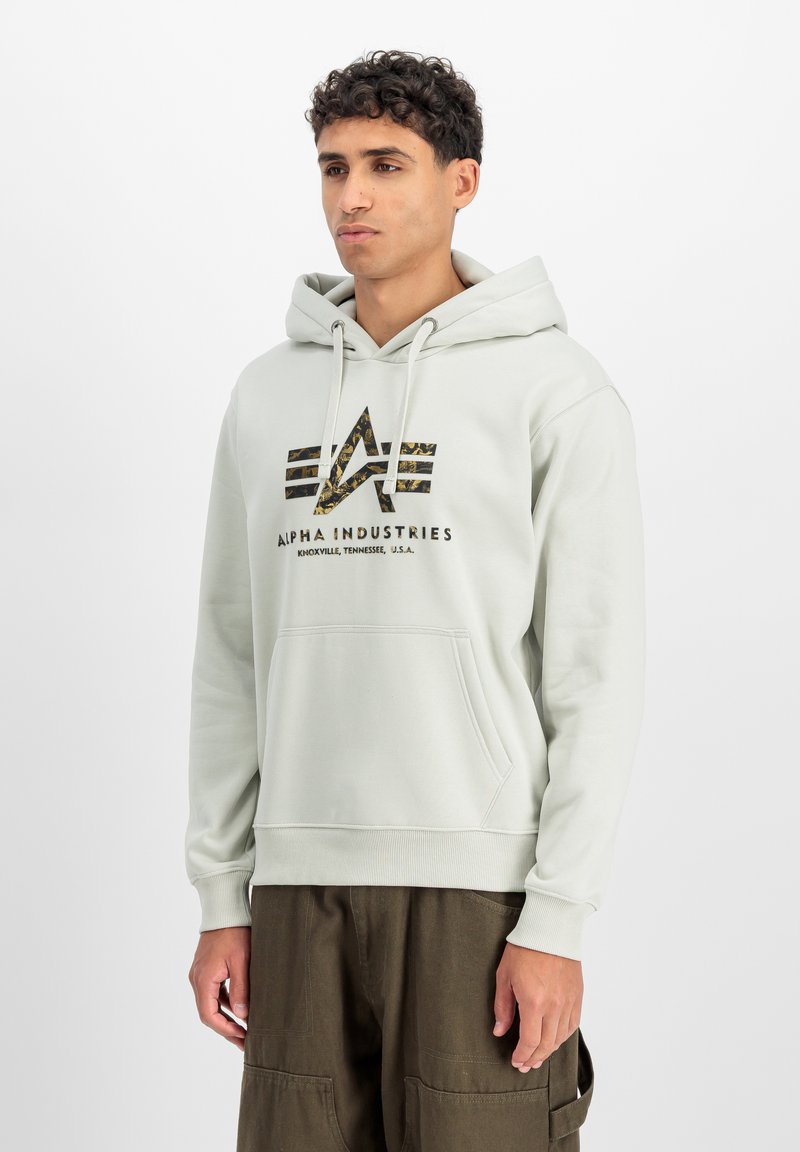 Sweat à capuche gris clair en matière douce, avec une poche frontale et un logo graphique noir et camouflage. Poignets et ourlet côtelés.