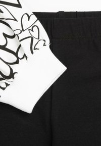 Manche blanche avec motifs de cœurs noirs et bord-côte assorti, associée à un tissu de ceinture élastique noir uni.