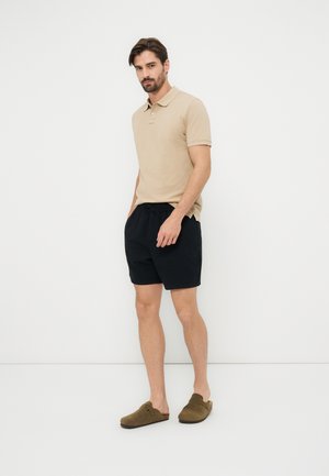 Homme debout sur fond blanc, portant un polo beige, un short noir et des chaussures marron sans lacets.