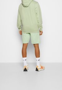 Felpa verde chiaro con cappuccio, pantaloni corti abbinati, calzini bianchi e sneakers con dettagli arancioni. Tessuto morbido, vestibilità comoda, design minimale.