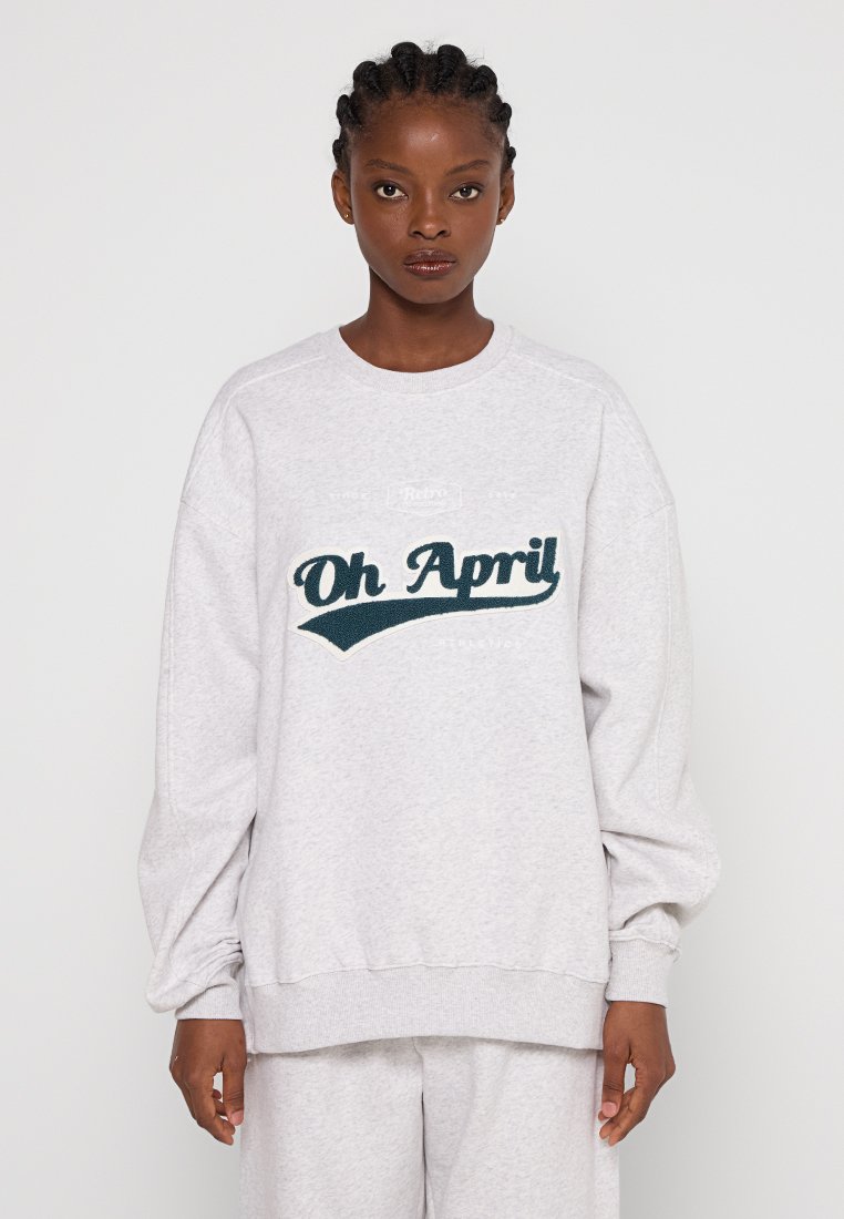 Junge Frau mit geflochtenem Haar, die einen hellgrauen, übergroßen Sweatshirt und Jogginghosen trägt. Auf dem Sweatshirt steht in dunkelgrüner Schrift "Oh April".