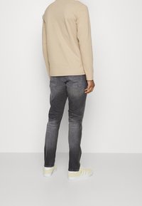 Mężczyzna w beżowej koszuli z długim rękawem, wyblakłych czarnych dżinsach i beżowych tenisówkach stojący tyłem do białej ściany.