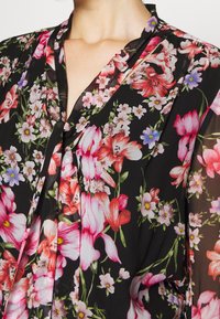 Kwiatowa bluzka z przezroczystego materiału z czarnym tłem, ozdobiona żywymi różowymi i białymi kwiatami, z subtelnym detalem plisy oraz dekoltem w serek.