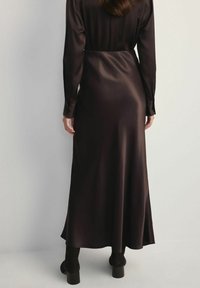 Kobieta ubrana w długą, ciemnobrązową satynową suknię i czarne botki na słupku, stojąca przodem do jasnej ściany.
