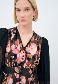 Mørk blomsterkjole med pink og brune nuancer, V-udskæring, sorte knapdetaljer og gennemsigtige puffærmer for ekstra tekstur.